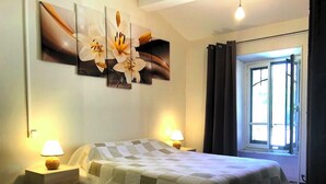 A bedroom with a checkered floor, a bed with a quilt, two bedside lamps, and a window with curtains. - The press house (minerve)