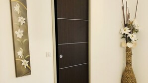 A hallway with a dark wooden door, a wall-mounted floral artwork, and a tall woven vase with white flowers.