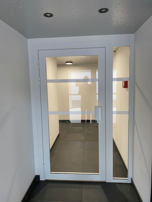 Een gang met een glazen deur naar een kamer. De vloer is betegeld en het plafond heeft inbouwspots.