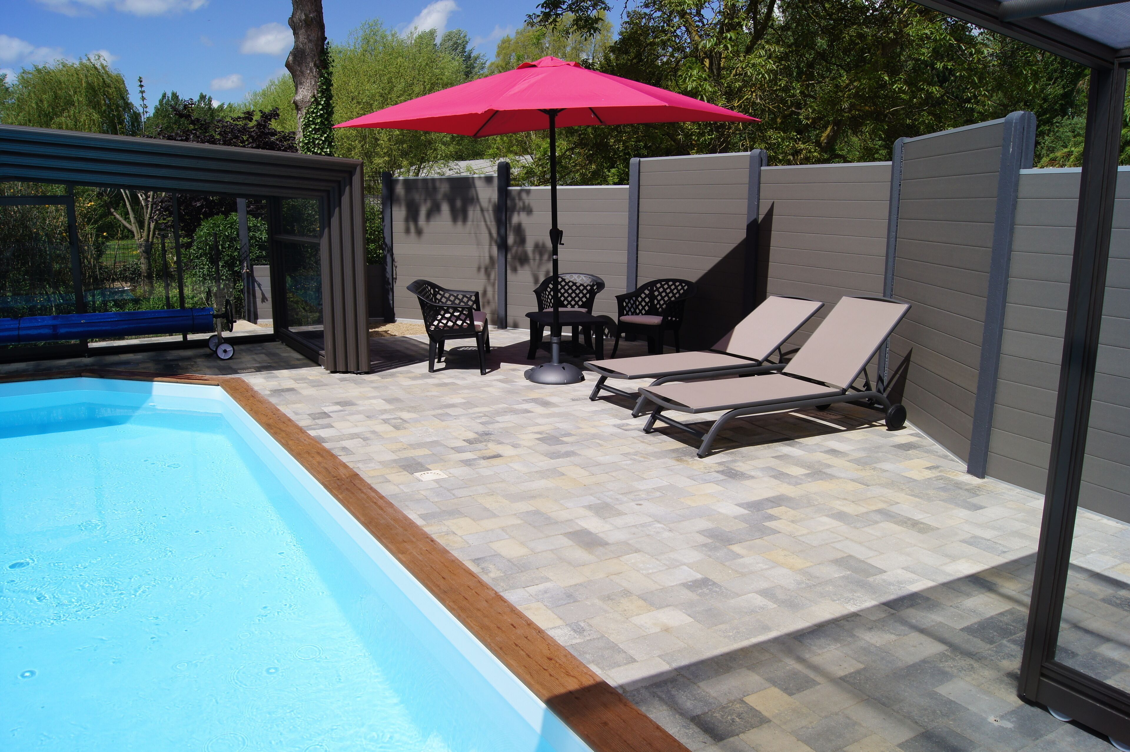 A poolside area with a blue pool, lounge chairs, a red umbrella, and a covered seating area.