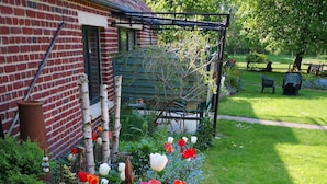 Un jardin où l’on trouve une grande variété de fleurs et de plantes, un bâtiment en briques et un banc en bois.