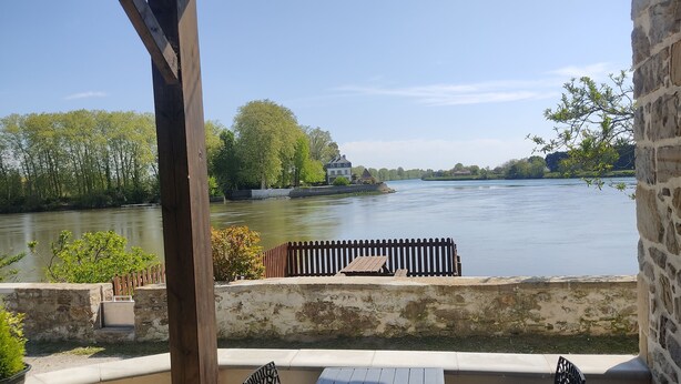 A riverside patio with a table and chairs, stone walls, and a view of a river and trees.