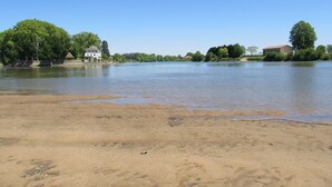A sandy beach with a calm river, surrounded by trees and buildings.