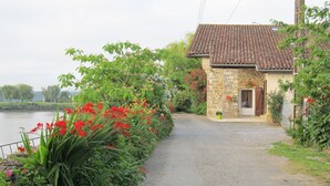 A rural road leading to a stone house with a tiled roof, surrounded by lush greenery and flowering plants.