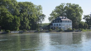 A large, multi-story house by a calm river, surrounded by lush greenery.