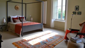 A four-poster bed with a red blanket, a patterned rug, a window with a view of greenery, and a chair with a hat. - Charming guest rooms (Lessac)