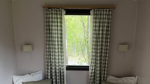 A small, single-room cabin with two beds, a window with checkered curtains, and a view of greenery outside. - Basecamp Birtavarre (Kåfjord)
