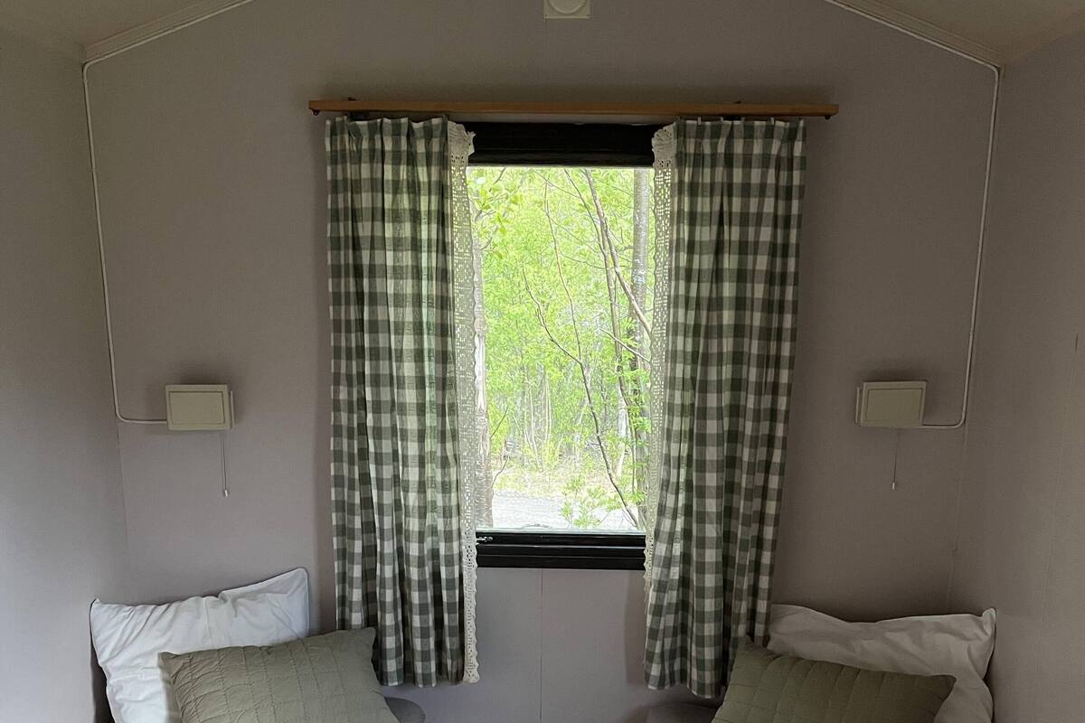 Un piccolo chalet con una stanza, due letti, una finestra con tende a quadretti e vista sul verde.