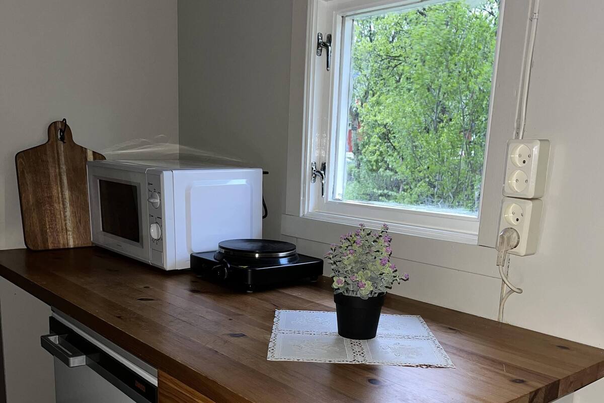 Una cucina con un forno a microonde, un piccolo elettrodomestico nero, un tagliere di legno, una pianta in vaso e una finestra con vista sul verde.