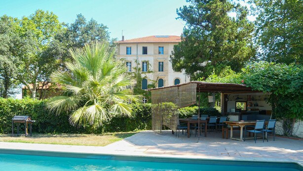 A poolside area with a covered seating area, a barbecue grill, and a building with a tiled roof in the background. - Chateau in magnificent park with swimming pool (Thuir)