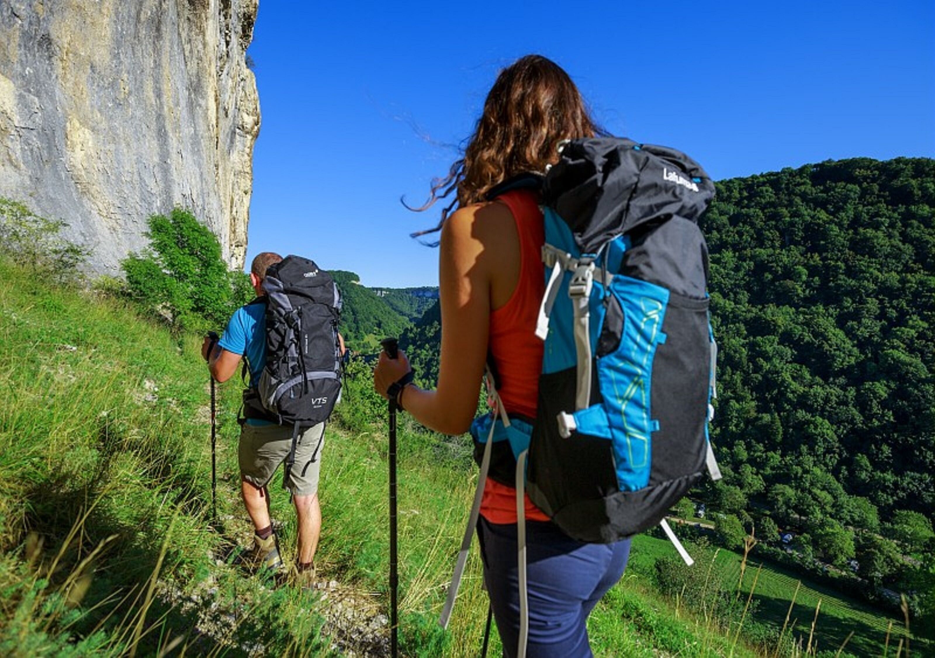 Two hikers with backpacks on a grassy trail with a cliff in the background.
