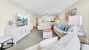 A spacious living room with a flat-screen TV, white furniture, and a view into the kitchen. - Award-Winning Resort Amenities | Seascape 219 (Isle of Palms)