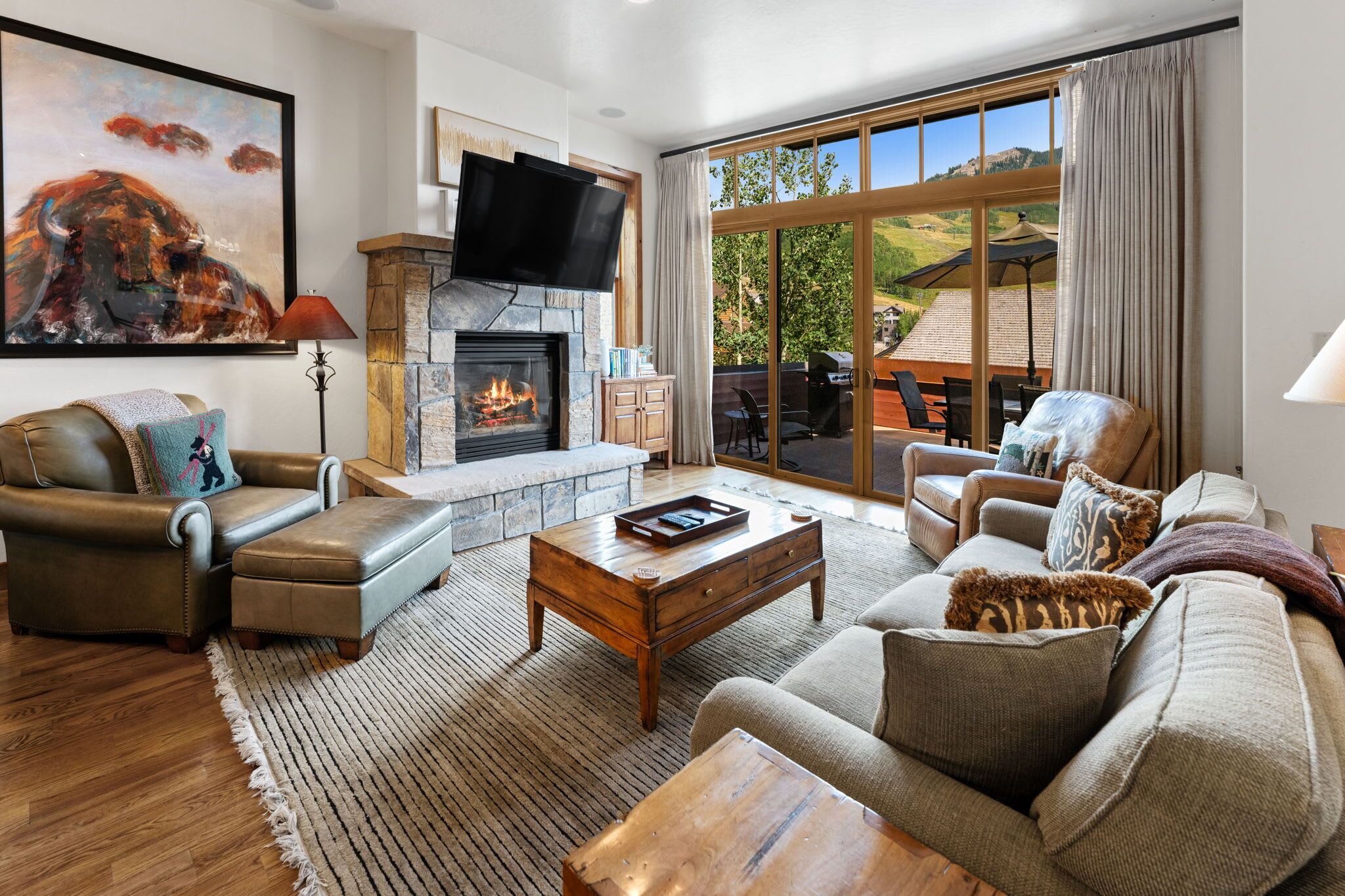 A spacious living room with a stone fireplace, leather furniture, and a large window offering a view of the outdoors.