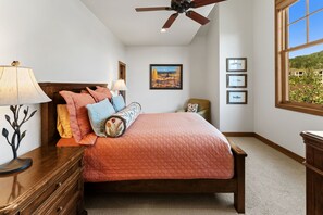 A bedroom with a bed, a nightstand, a lamp, a ceiling fan, a window with a view, and framed pictures on the wall.