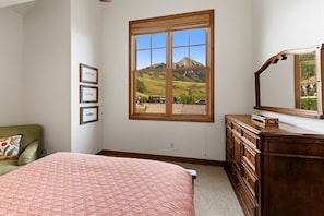 A bedroom with a large window, a bed, a dresser, and framed pictures on the wall. - Grand Villa (Crested Butte)
