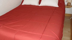A single bed with a red bedspread and two white pillows, a wooden nightstand with a lamp, and a power outlet on the wall. - Mountain apartment 6 people (Génos)