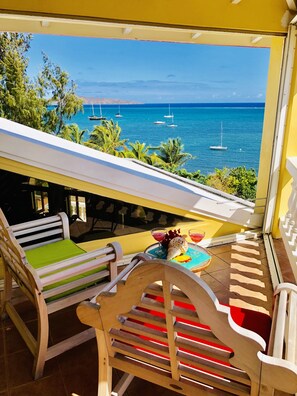 Un balcon avec vue sur la mer, des chaises en bois, une table avec des boissons et un coquillage décoratif.
