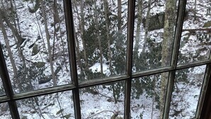 Ein Zimmer mit einem großen Fenster mit Blick auf einen verschneiten Wald. Im Inneren steht ein Tisch mit einer Kerze, Muscheln und einem verpackten Geschenk.