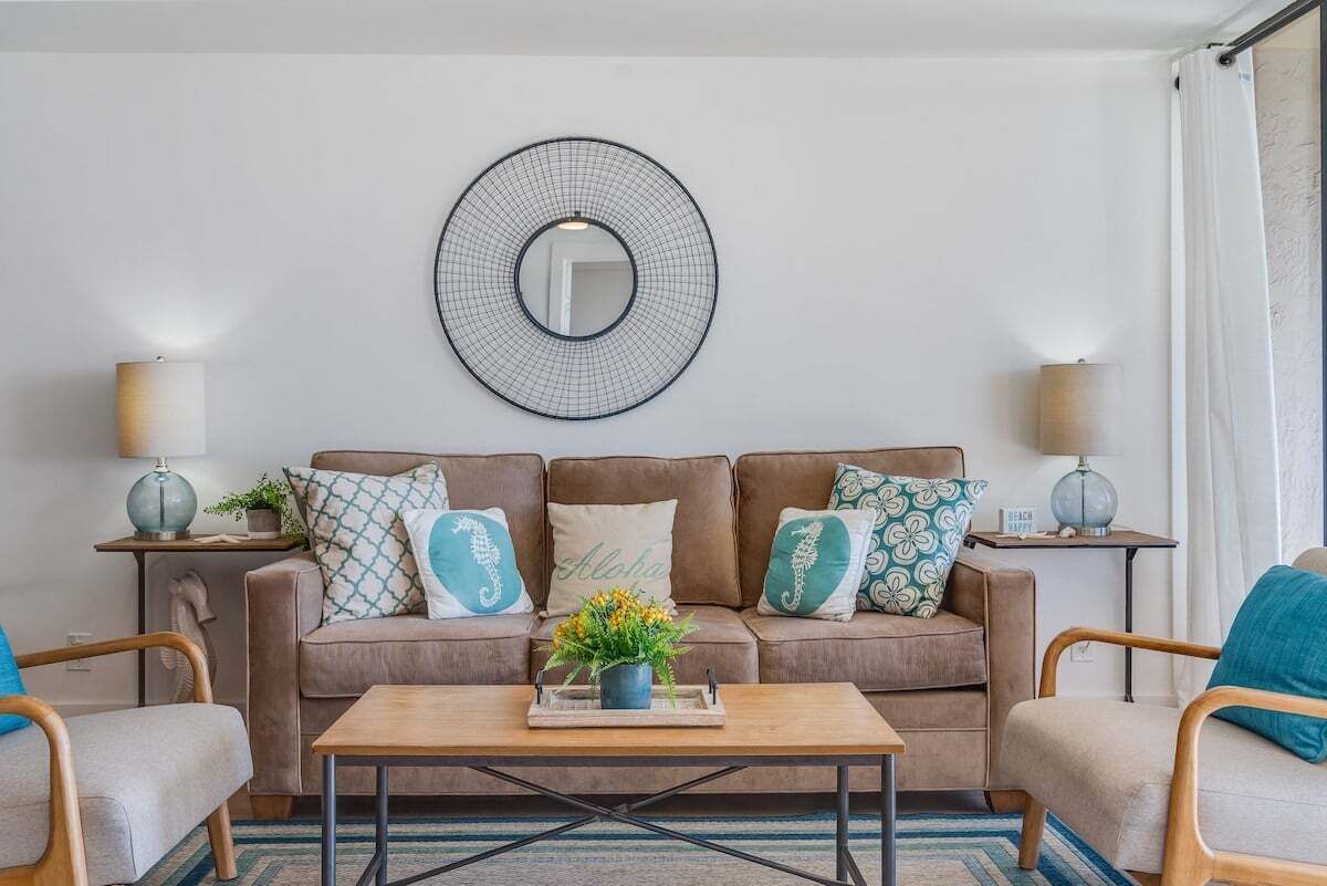 Un salon avec un canapé marron, deux tables d’appoint avec des lampes, une table basse avec un plateau et des fleurs, et un grand miroir rond accroché au mur.