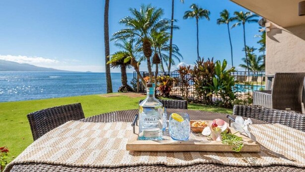 A table with a bottle of vodka, a glass of water, and a tray with snacks and a flower, set against a backdrop of a pool and palm trees. - SUMMER SALE! 2BD OceanFront Getaway w AC Pool - Lauloa 105 (Wailuku)