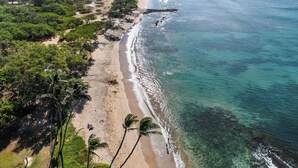 A coastal landscape with a beach, palm trees, and a distant industrial facility. - SUMMER SALE! 2BD OceanFront Getaway w AC Pool - Lauloa 105 (Wailuku)