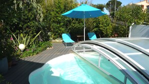 Een zwembad met een gebogen glazen overkapping, omringd door een houten terras en een blauwe parasol.
