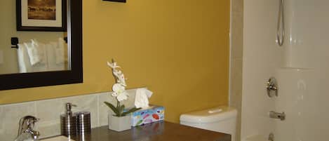 Une salle de bain avec un lavabo, des toilettes et une douche. Un tableau encadré est accroché au mur et un vase avec des fleurs est posé sur le comptoir.