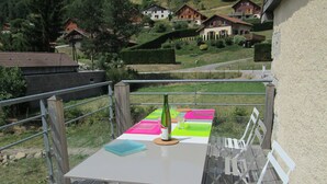 A table with a bottle, glasses, and a placemat on a wooden deck with white chairs. - Duplex "La Ferme de Paul" 6 Pers. + baby - Altitude 800 m'at 2 km Ski slopes (La Bresse)