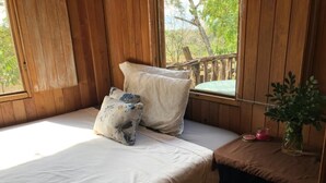 A wooden cabin room with a bed, pillows, a small table with a plant, and a window with a view of trees.
