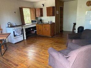 A compact kitchen with wooden cabinets, a microwave, a gas stove, and a sink. There is a wooden dining table and a purple sofa in the living area.