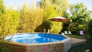 Una piscina de fusta amb una bassa integrada i una ombrelles, envoltada de vegetació abundant.