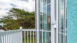 Een balkon met een witte reling, een glazen deur en zicht op een boom en gebouw op de achtergrond.
