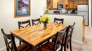 A dining area with a wooden table and chairs, a vase with yellow flowers, and a kitchen in the background. - Terracehouse 2BR Ski Getaway, Steps to Lifts, Fireplace + Hot Tub (Snowmass Village)