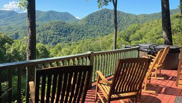 A deck with wooden chairs and a grill, overlooking a forested mountain range.