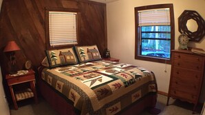 A bedroom with a wooden headboard, two windows with blinds, a bed with a quilt, a dresser, and a lamp.