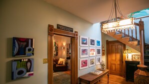 A hallway with a staircase, wooden walls, and a chandelier. There are framed pictures on the wall and a table with a lamp.