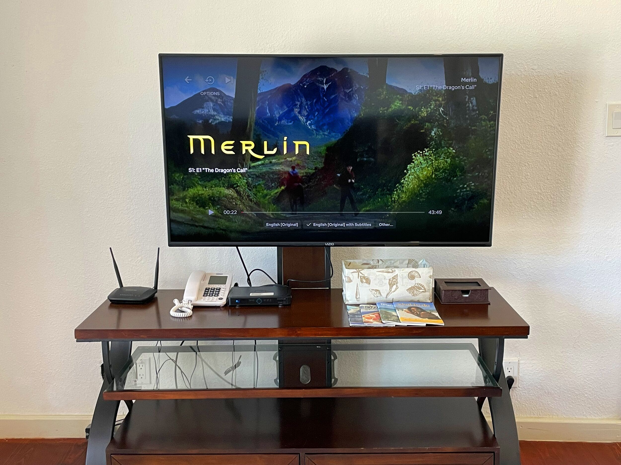 Un televisor sobre un soporte de madera, con un control remoto, un teléfono y algunas revistas.