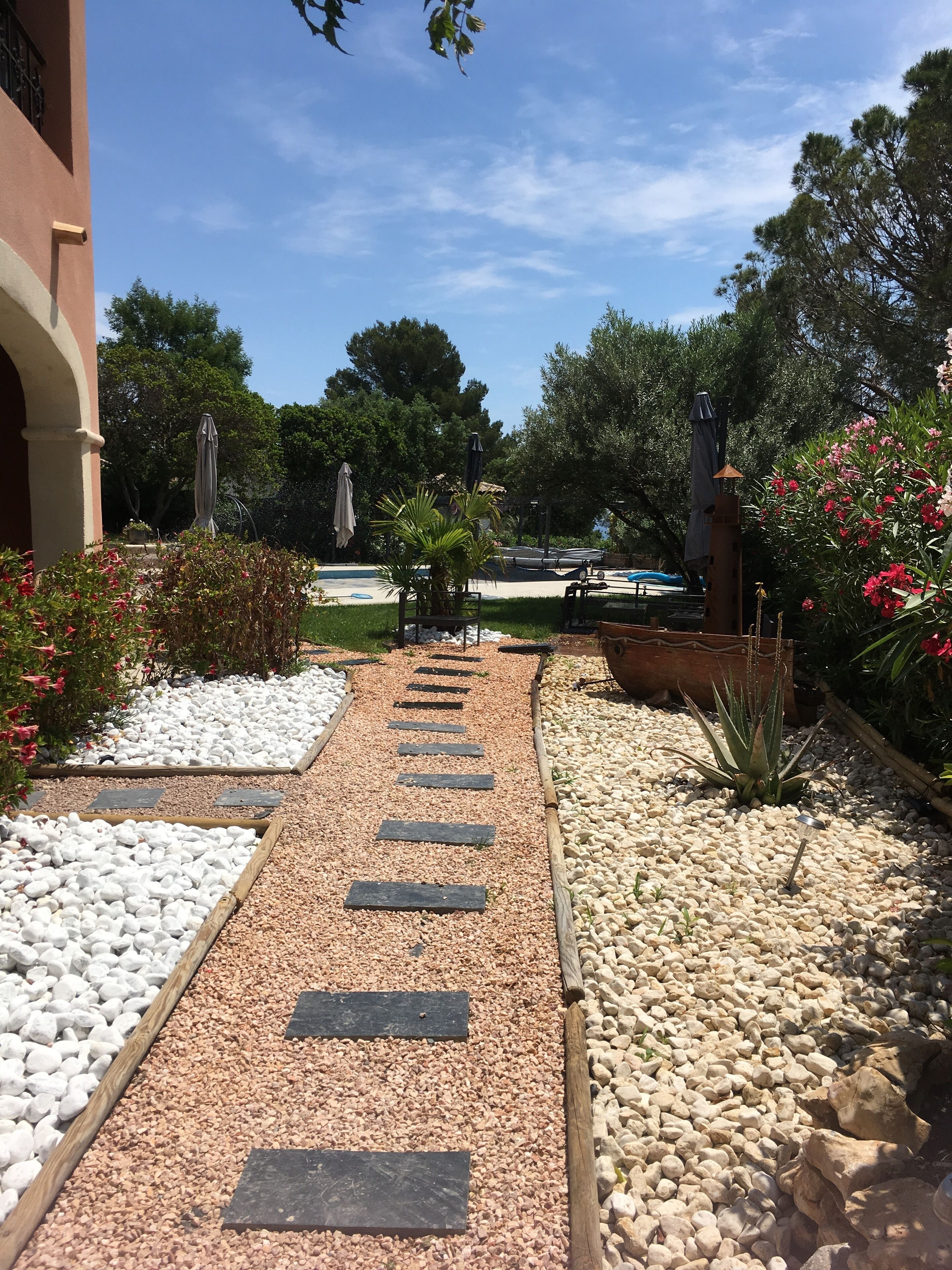 A pathway lined with white and beige pebbles, leading to a pool area with umbrellas and a statue.