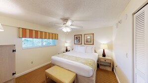 A hotel room with a bed, a bedside table with a lamp, a ceiling fan, a window with blinds, and a door with a window shutter. - Resort Sixty-Six - 1 Bedroom (Holmes Beach)
