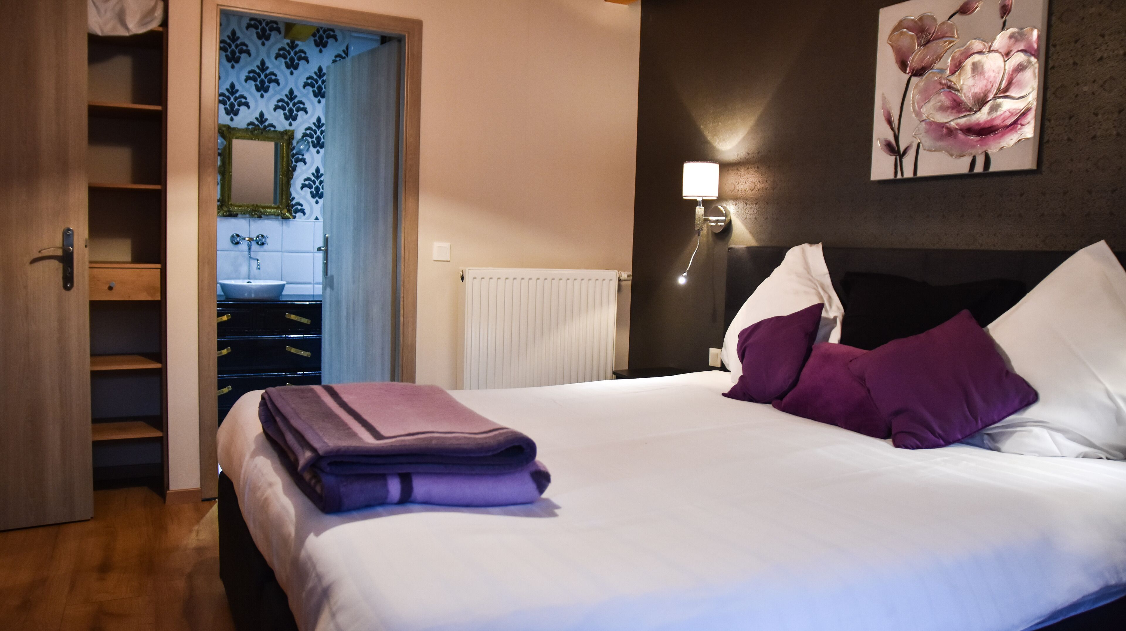 A hotel room with a bed, a nightstand with a lamp, a bathroom with a sink and mirror, and a wardrobe.