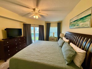 A hotel room with a bed, a television on a dresser, a ceiling fan, and a painting on the wall. - Ocean Oasis (Volusia County)