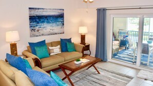A living room with a sofa, two side tables with lamps, a coffee table, a painting of a beach, and a sliding glass door leading to an outdoor area with a patio set. - Ocean Oasis (Volusia County)