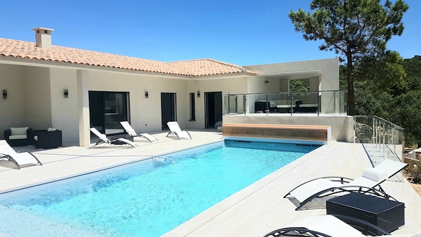 Uma área moderna à beira da piscina com espreguiçadeiras, piscina e um edifício com telhado cerâmico.
