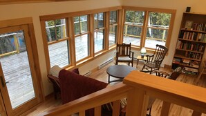 A cozy room with a wooden floor, a red sofa, a small table, and a bookshelf filled with books. Large windows offer a view of a wooden deck outside. - Lakefront cottage near Acadia and Bar Harbor (Sullivan)