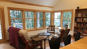 A cozy living room with a sofa, chairs, a coffee table, and a bookshelf. Large windows offer a view of trees outside. - Lakefront cottage near Acadia and Bar Harbor (Sullivan)