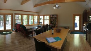A spacious living area with a wooden floor, a dining table, a bookshelf, and large windows offering a view of the outdoors. - Lakefront cottage near Acadia and Bar Harbor (Sullivan)