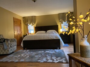 Un dormitorio con una cama grande, cabecera de madera, mesita de noche con lámpara, una silla, un jarrón decorativo y un tapete estampado.