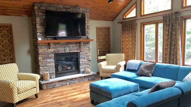 A living room with a stone fireplace, a flat-screen TV mounted above it, a blue sofa, and patterned chairs.