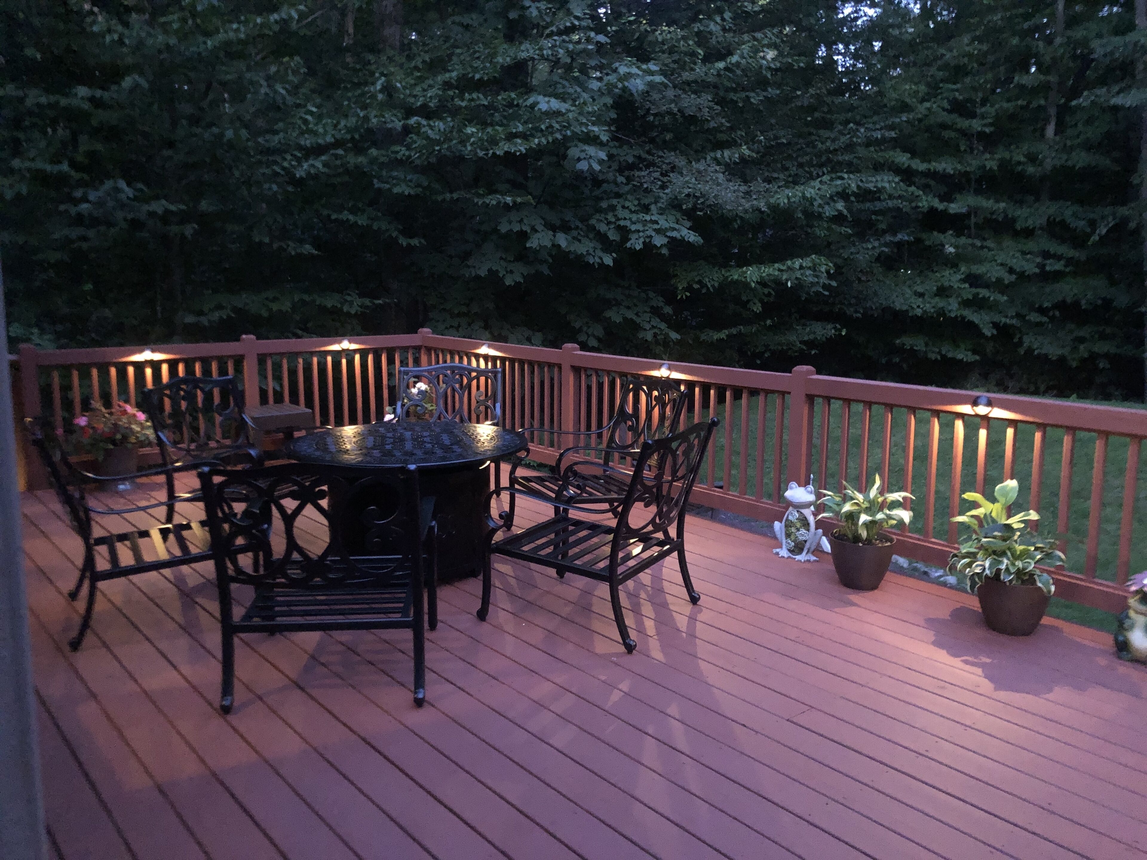 A wooden deck with a round table and chairs, surrounded by potted plants and a small frog statue.