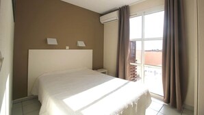 Ein Hotelzimmer mit einem Bett, einem Nachttisch, einer Wand-Klimaanlage und einem Vorhangfenster.